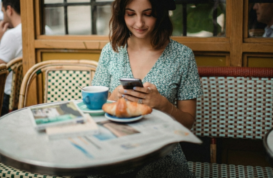 Phone Notifications Are Hijacking Your Attention. A young woman wearing a dark beret and a green floral dress looks down at her smartphone while sitting at an outdoor cafe. In the foreground, a blue coffee cup and a fresh croissant sit on a marble-topped table, with blurry cafe chairs and other patrons in the background.