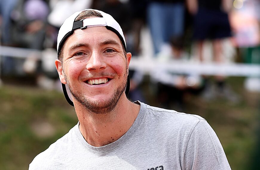 Jan-Lennard Struff hits a forehand during an ATP tennis match, wearing a white cap and black-and-yellow kit with a blurred stadium background